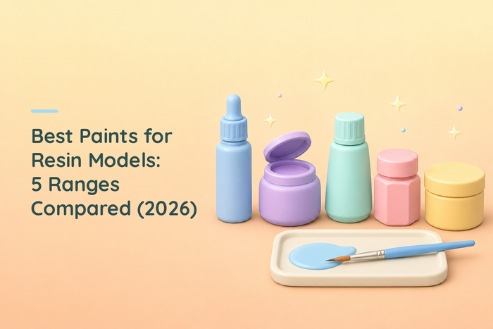 Five differently shaped hobby paint containers in a row in soft pastel colours, one open, with a palette tray holding a small pool of blue paint and a fine brush resting across it